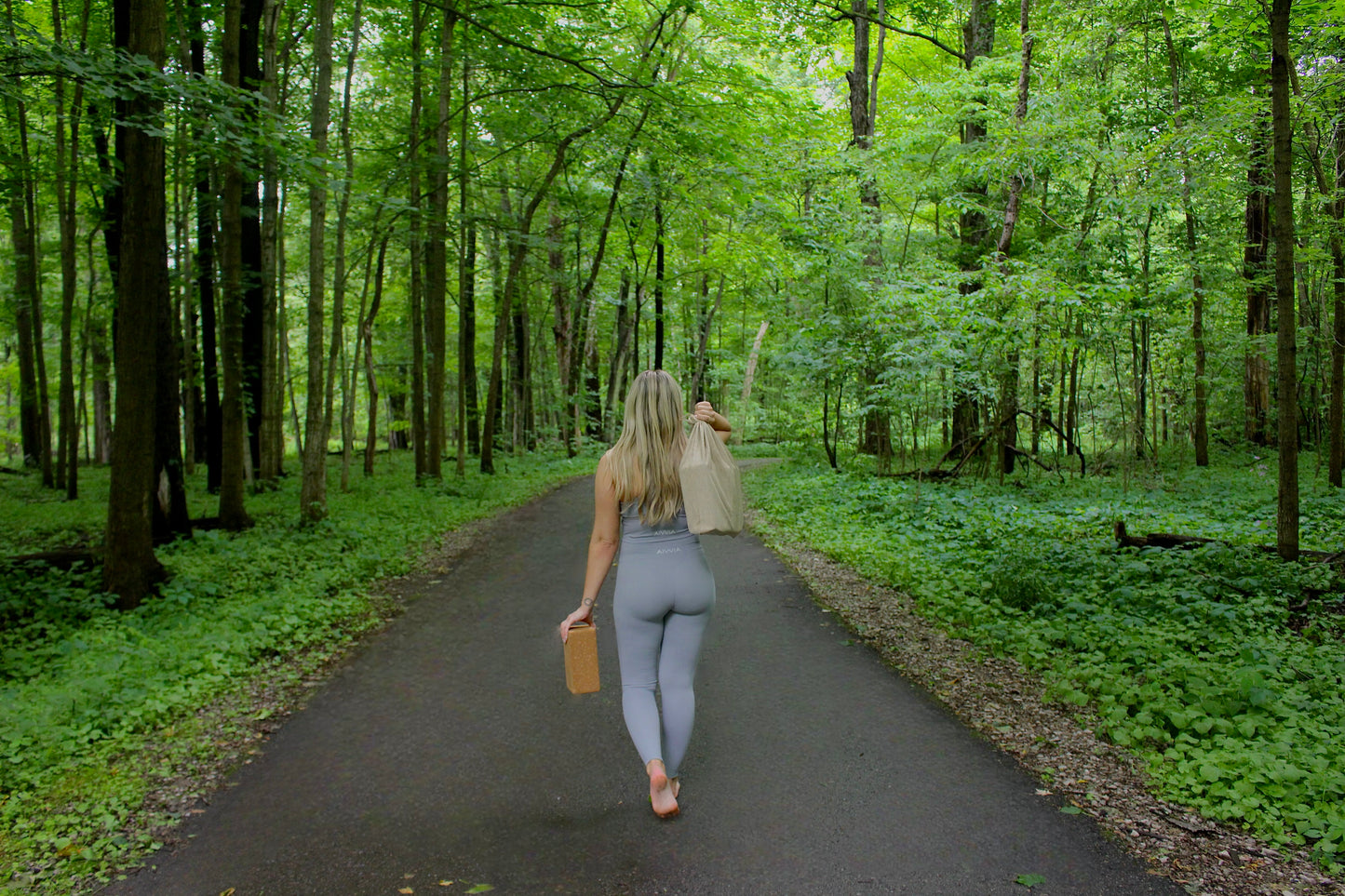 Woman walking down a forest path carrying cork yoga blocks and a rolled mat.