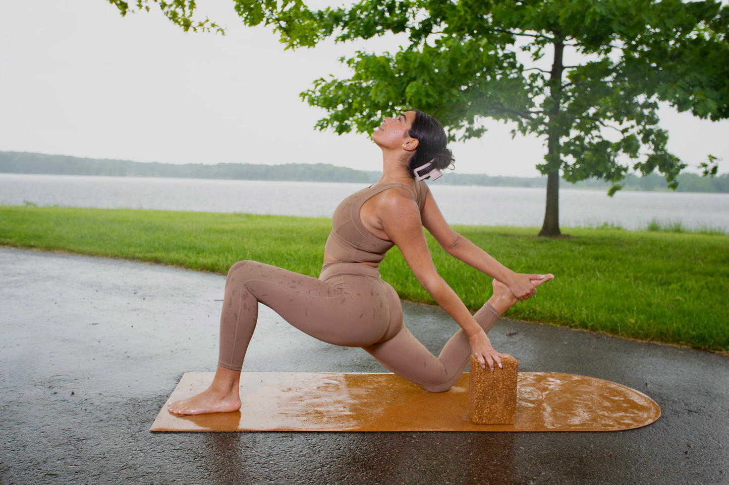 Woman performing a deep lunge stretch on a cork yoga mat, using a cork yoga block under her hand for balance during an outdoor session by the lake.