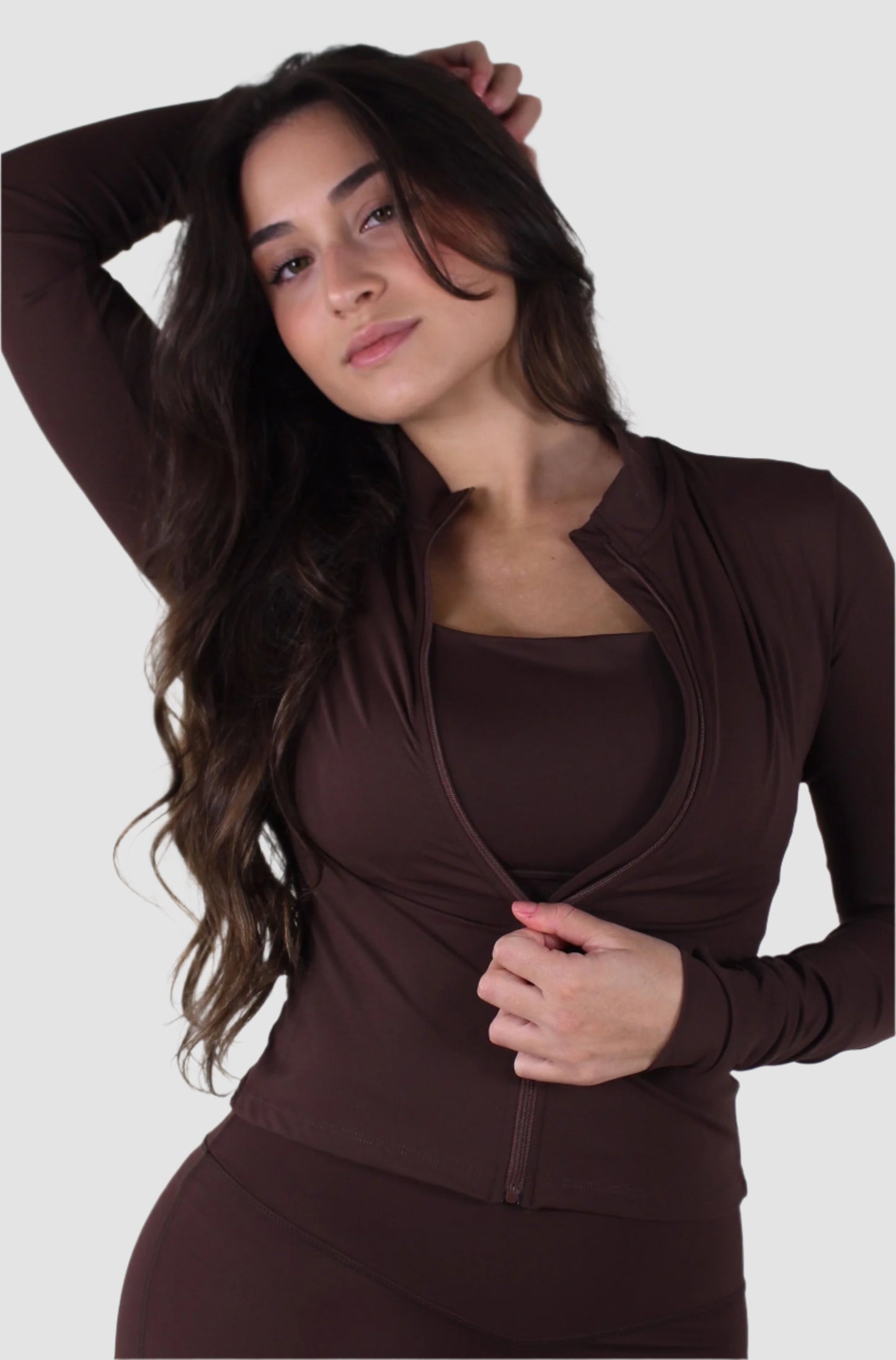 Woman posing in brown Aivvia Athletics long-sleeve fitted jacket, zipped halfway over matching set.