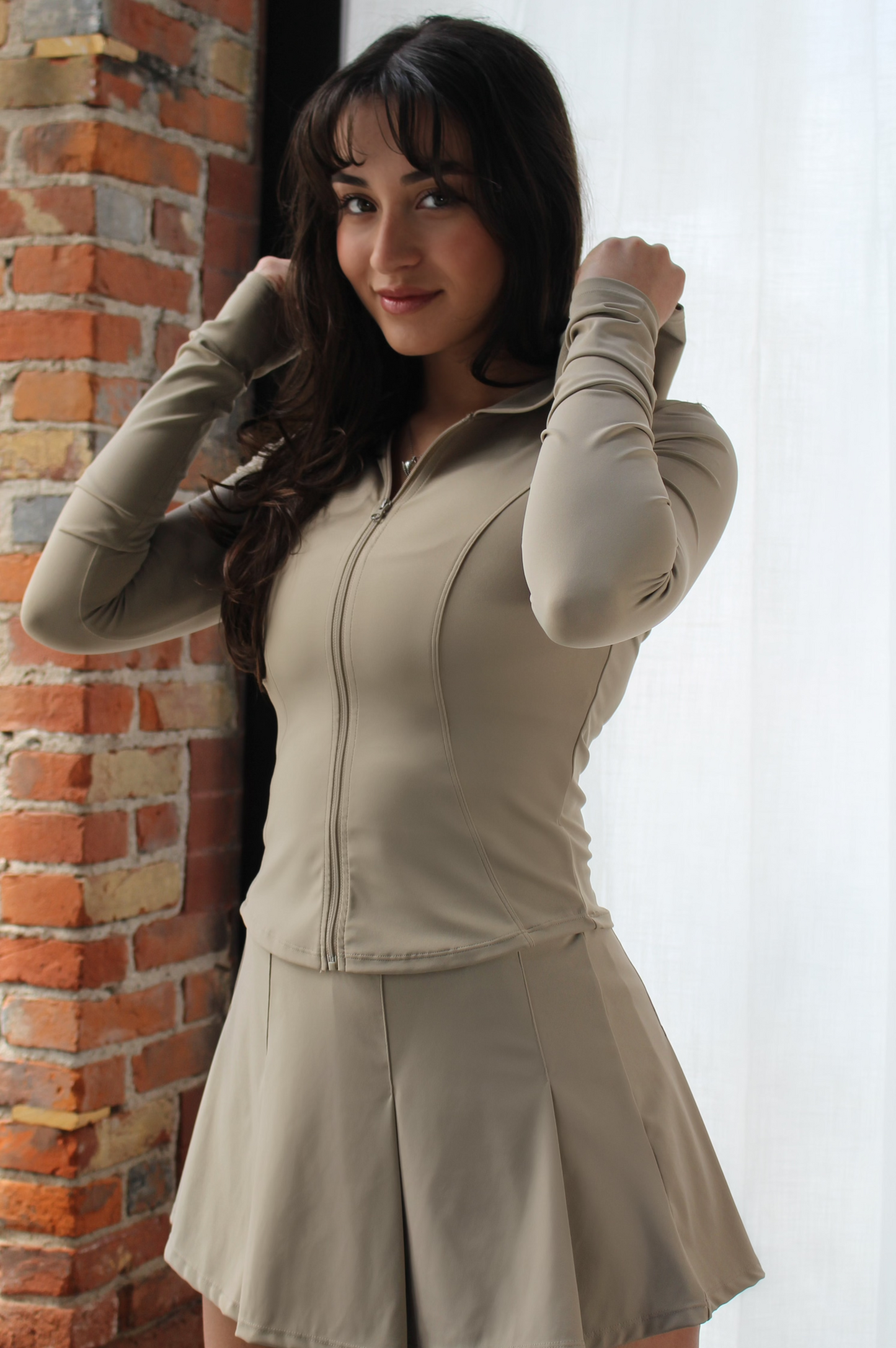Woman standing in Aivvia Athletics beige hoodie and pleated tennis-style skirt — soft and fitted activewear for workouts, recovery, and casual wear.
