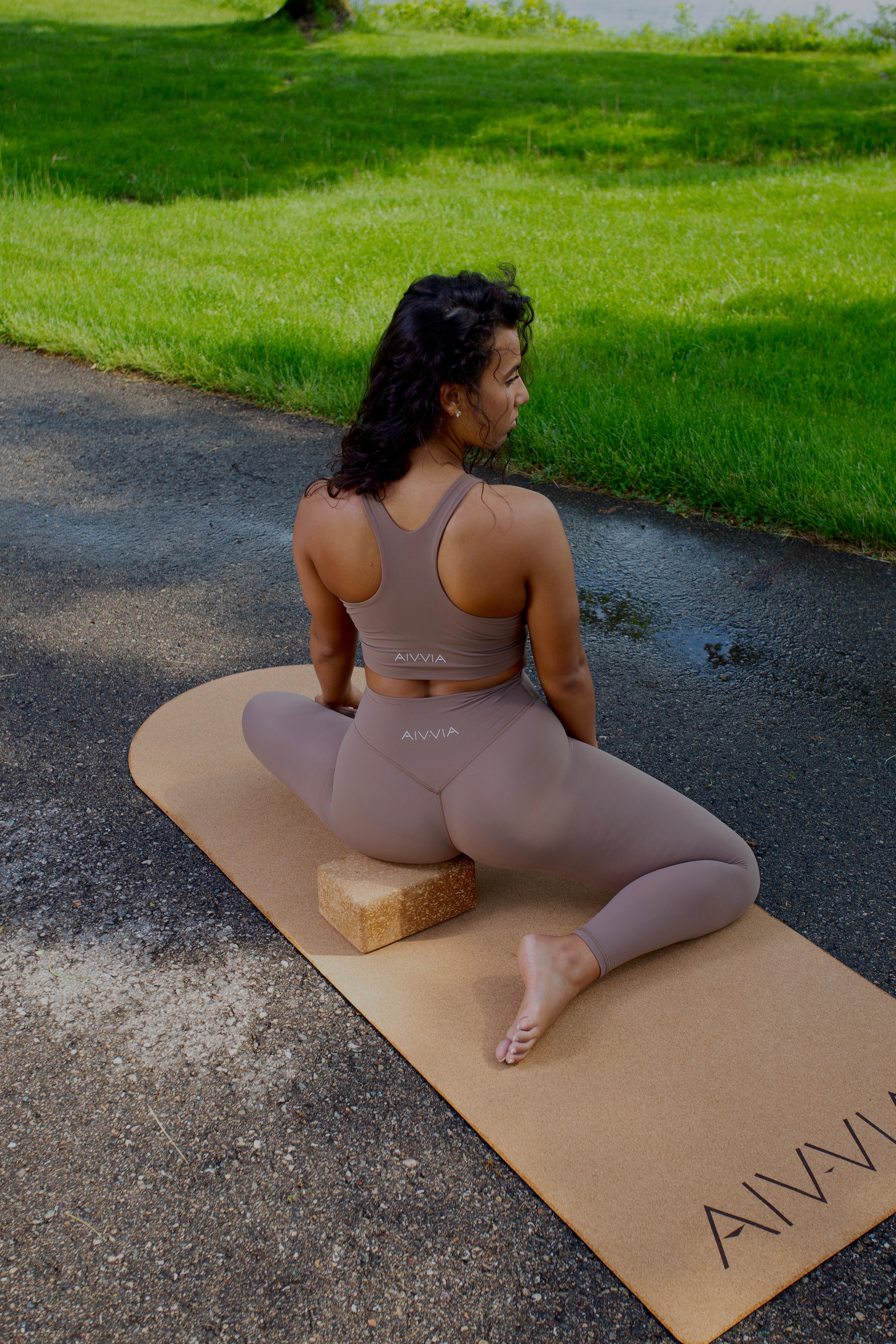 Woman seated cross-legged on a cork yoga mat in the sun, with arms resting behind her.