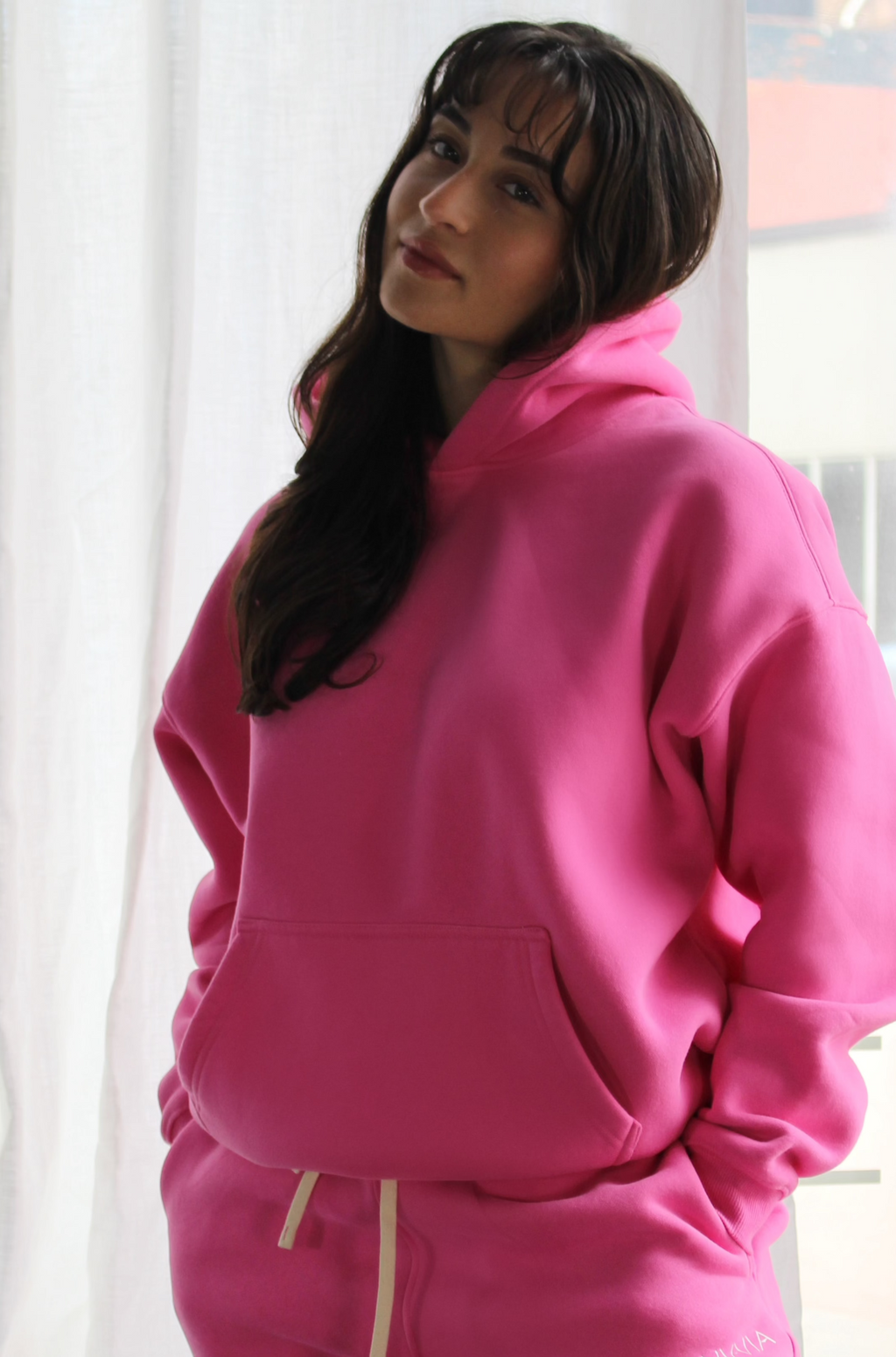 Bold pink Aivvia Athletics hoodie in a relaxed pose; cozy fleece material, drop-shoulder design, and modern loungewear style.