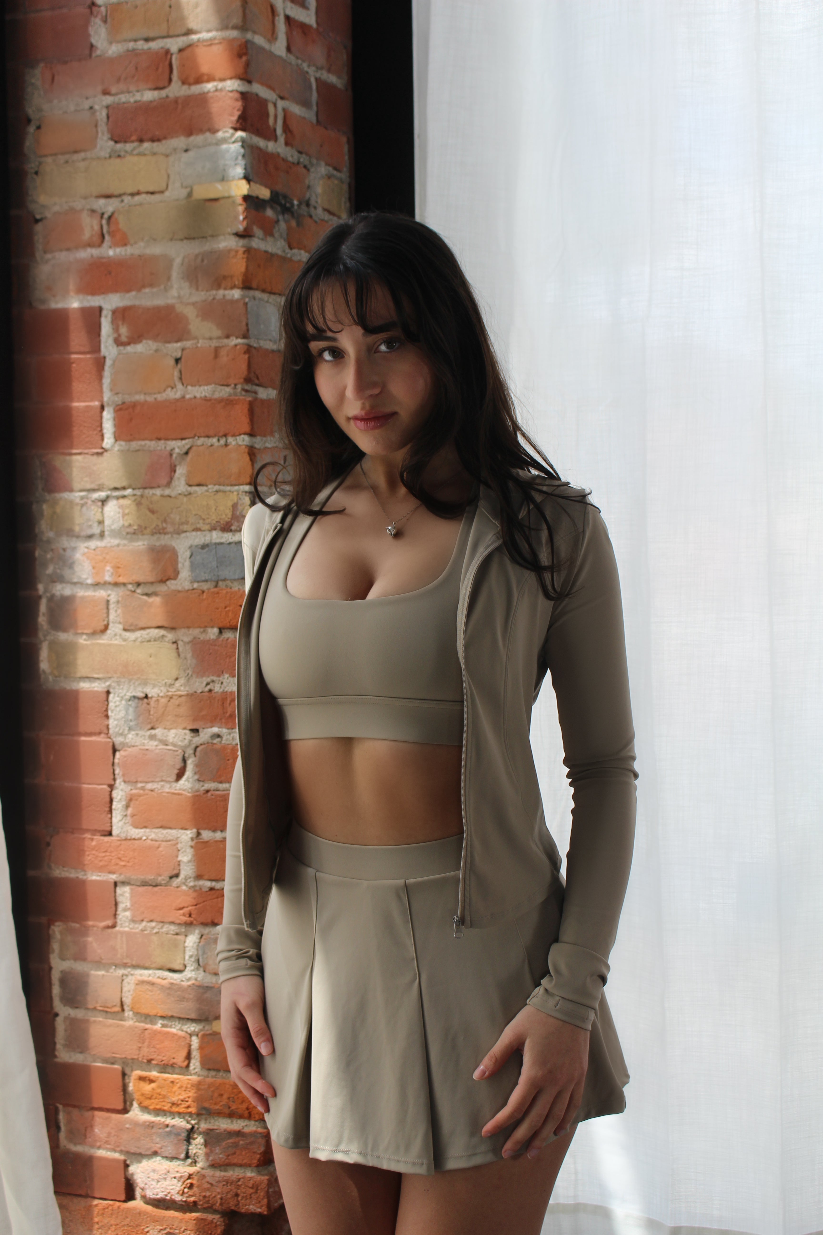 Woman wearing Aivvia Athletics beige pleated skirt, sports bra, and matching long-sleeve top — full activewear ensemble perfect for layering and seasonal versatility.