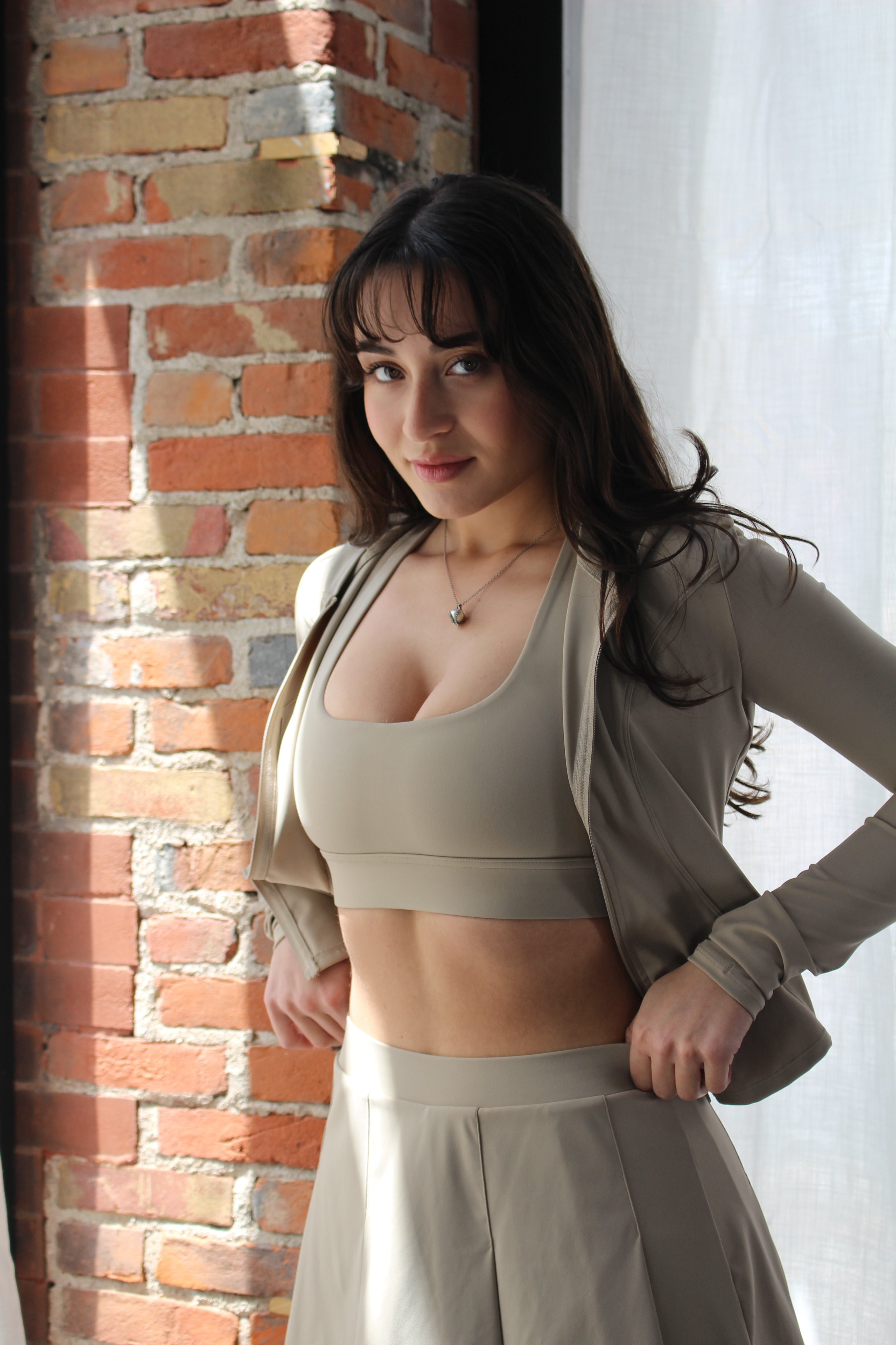 Woman adjusting Aivvia Athletics beige zip-up jacket over sports bra and skirt — sculpted, high-performance workout jacket for cool-downs and warmups.