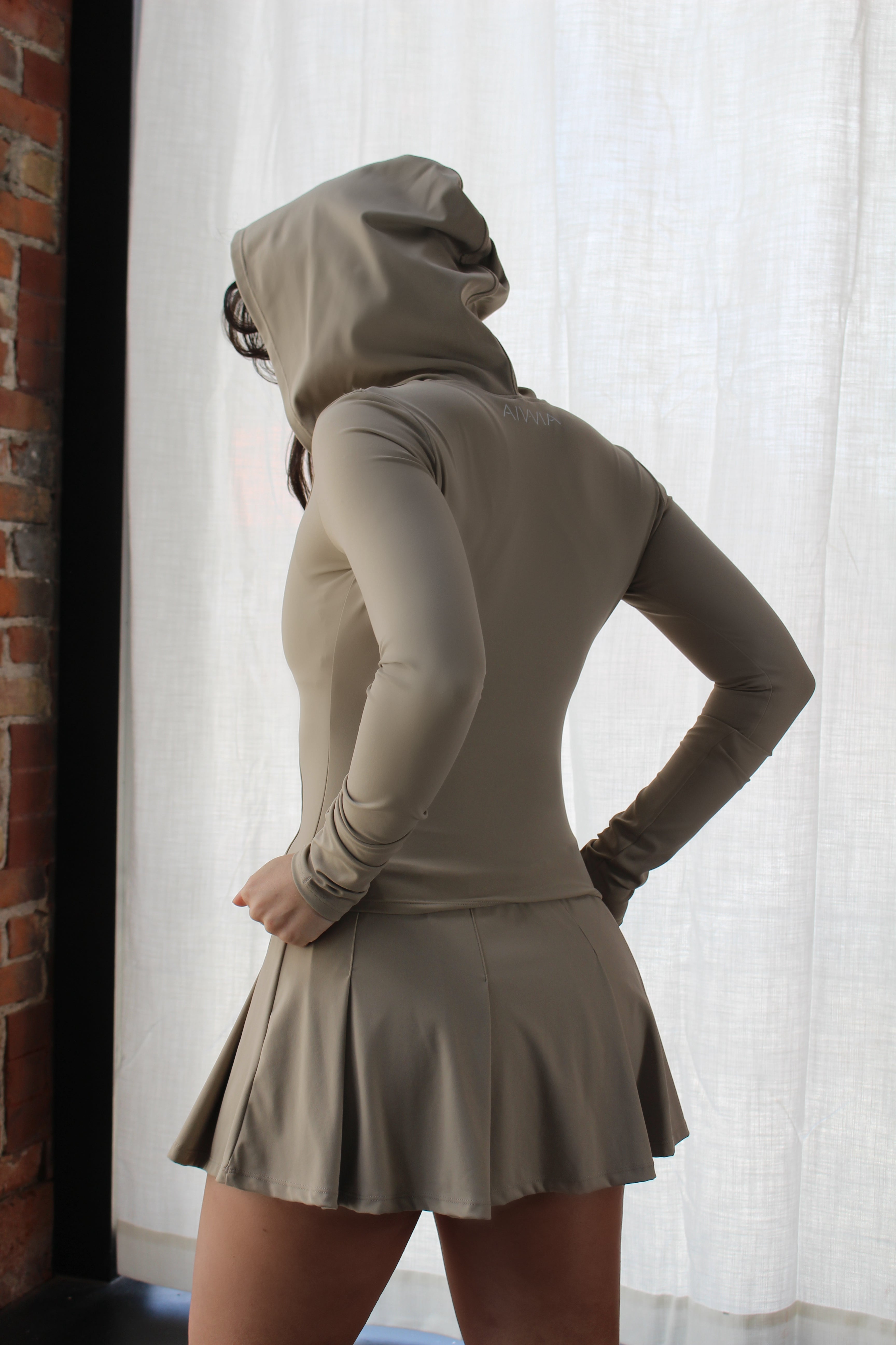 Side profile of woman in Aivvia Athletics beige hoodie and pleated active skirt — functional, feminine activewear look with streamlined hooded jacket.
