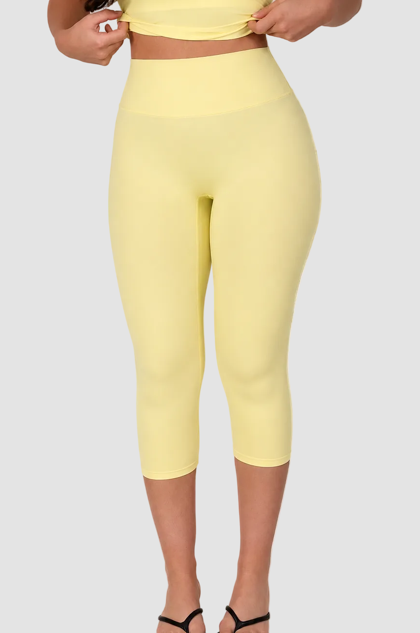 A model wearing high-waisted, form-fitting yellow capri leggings and a matching yellow crop top, paired with black flip-flop sandals, against a plain white studio background.
